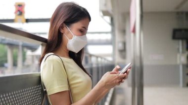 Yüz maskesi takan güzel bir Asyalı kadın elinde akıllı bir telefonla platformda tren bekliyor. Trafik ve sermaye taşımacılığı kavramı. İş çıkış saatinde seyahat etmek kolaydır.