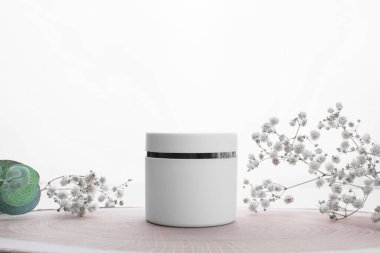 Tahta bir kürsüde, bitkilerle, çiçeklerle dolu beyaz bir krema kavanozunun kopyası. Kapalı beyaz nemlendirici kavanozu. Beyaz nemlendirici kavanozu