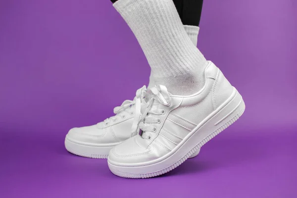 Women\'s legs in stylish white sneakers. Sneakers on legs on a purple background