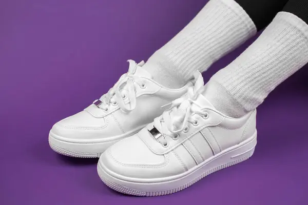 Women\'s legs in stylish white sneakers. Sneakers on legs on a purple background