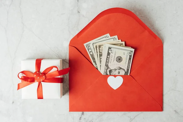 stock image Red paper envelope with money dollars bills. Flat lay of gray working table background with Valentine gift, letter, heart shape. Top view, mock up greeting card. Valentines Day budget