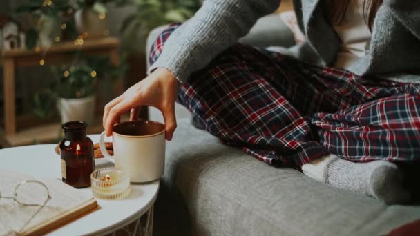 舒适的女人腿 穿着针织的冬季保暖袜子和格格的睡衣 在杯子里喝着热可可或咖啡 坐在家里的沙发上 秋天有蜡烛 装饰品 饼干和室内植物 — 图库视频影像