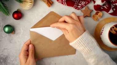 Kadın kazak giymiş, boş zarf içinde tebrik kartı yazıyor. Eko doğal Noel süslemesi, kahve fincanı, köknar dalı ile bembeyaz ahşap arka plan. Üst görünüm