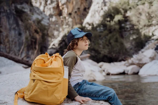 School boy with yellow backpack sits on a riverside rock in the canyon with mountain cliffs in the background. Kid child taking a rest on a boulder near mountain river.