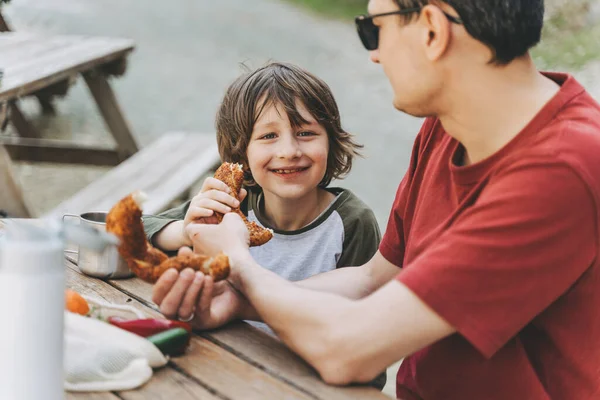 Close-up view of father hugs his school boy son on a family picnic. Child kid and his dad taking a rest and enjoying a picnic while hiking. Boy smiles and bites the bread donut on a picnic.