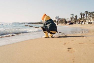 Boy in yellow rubber boots playing with stick and sand at the beach. School kid touching water at autumn winter sea. Child having fun with waves at the shore. Spring Holiday vacation concept.