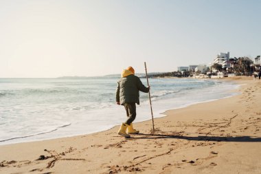 Boy in yellow rubber boots playing with stick and sand at the beach. School kid touching water at autumn winter sea. Child having fun with waves at the shore. Spring Holiday vacation concept.