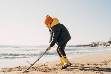 Boy in yellow rubber boots playing with stick and sand at the beach. School kid touching water at autumn winter sea. Child having fun with waves at the shore. Spring Holiday vacation concept.