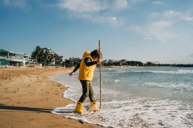 Boy in yellow rubber boots playing with stick and sand at the beach. School kid touching water at autumn winter sea. Child having fun with waves at the shore. Spring Holiday vacation concept.