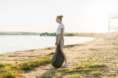 Çantalı ve eldivenli genç bir gönüllü çöpleri, plastik şişeleri topluyor, sahili denizle temizliyor. Çöp toplayan kadın. Çevresel ekoloji kirliliği kavramı. Dünya Günü.