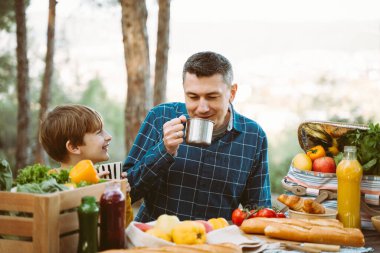 Babası ve okul çocuğu ormanda piknik yapıyorlar sebzeler, meyve suyu, kahve ve kruvasanlarla. Ekmek baget ile çevrili taze organik sebzelerle dolu tahta sandık.