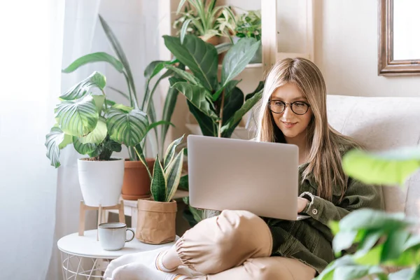 Freelance woman in glasses typing at laptop and working from home office. Happy girl sitting on couch in living room with plants in cozy urban jungle home. Distance learning online education and work.