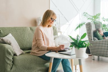 Yetişkin bir kadın dizüstü bilgisayarında online bankacılık yatırımları yaparken not defterine bir not yazar. Üst düzey bir bayan ev ofisinde dizüstü bilgisayar kullanıyor. Boşluk modelini kopyala.