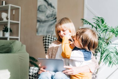 Son sınıf öğrencisi bir kadın ve oğlu tabletle oynuyor. Gülümseyen kadın ve torunu internet üzerinden alışveriş yapmak için dijital cihaz kullanıyorlar..