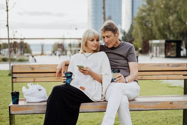 stock image An adult mature happy couple in love sitting on bench with phone outdoors in park. A blonde caucasian man and woman spend time together and drinking coffee. Senior wife and husband walking outside