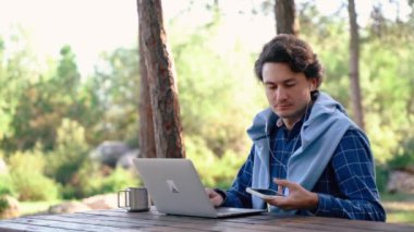 Bilgisayarlı ve cep telefonlu bir adam doğa parkında oturuyor. Bilgisayar yazma, mesaj yazma, blog yazma, dağlarda gezme konusunda serbest çalışan biri. Serbest çalışma, uzaklık çalışma konsepti.