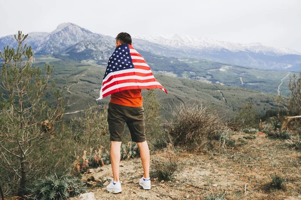 stock image Young man standing on rock cliff with the US flag on his back while looking at mountains in background. Male traveller wearing American flag standing on mountain top. 4 fourth July Independence Day.