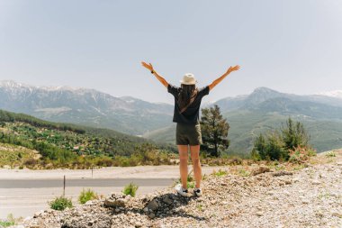 Young woman in hat standing on Mountain View. Raised hands on mountain background.