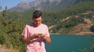 Man texting online on mobile phone of mountains lake background. Traveler male chat messages on cellphone on the blue lake outdoors travel adventure vacation. Using smartphone