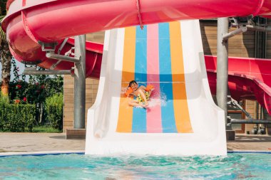 Su parkında güneşli bir günde su kaydırağına düştükten sonra havuzda kayarken gülen çocuk. Sıcak havalarda ferahlatıcı bir çocuk, aktif tatil ve sağlıklı bir yaşam tarzı. Mutlu yaz..