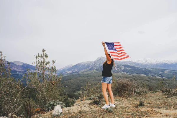 Young woman standing on a rock cliff and waving the US flag while looking at mountains in the background. Girl traveller waving American flag standing on mountain top. 4 fourth July Independence Day.