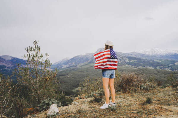 Девушка, стоящая на скале с флагом США на спине и смотрящая на горы на заднем плане. Женщина-путешественница с американским флагом на вершине горы. 4 июля День Независимости