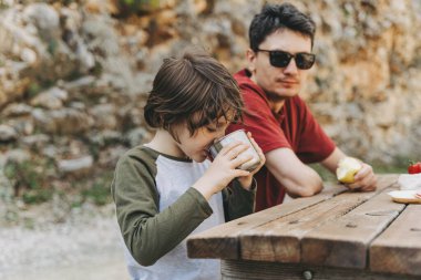 Babası ve oğlunun dağlarda aile pikniği yaparken çekilmiş yakın görüntüsü. Çocuk ve babası dağlarda yürüyüş yaparken dinlenip pikniğin tadını çıkarıyorlar..
