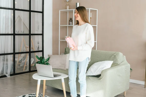 stock image Smiling young woman girl standing near her laptop with the notepad notebook in her hands. Modern home office decor.