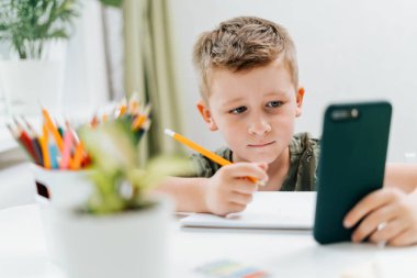 Okullu çocuk evde cep telefonuyla ders çalışıyor ve ödev yapıyor. Okula dönelim. Cep telefonu, kalem ve eğitim kitapları olan beyaz bir çocuk. Uzaklık öğrenimi çevrimiçi eğitim.
