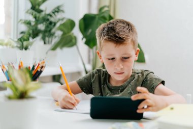 Okullu çocuk evde cep telefonuyla ders çalışıyor ve ödev yapıyor. Okula dönelim. Cep telefonu, kalem ve eğitim kitapları olan beyaz bir çocuk. Uzaklık öğrenimi çevrimiçi eğitim.