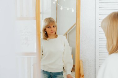 Beautiful smiling mature senior caucasian woman with blonde hair standing near mirror and looking at reflection at home.