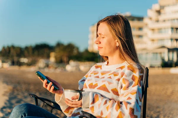 Genç ve güzel bir kadın elinde akıllı telefonuyla mesajlaşırken arka planda deniz dalgalarıyla kış sahilinde kahve içiyor. Sonbahar deniz kıyısında çay içerken cep telefonu kullanan sevimli kız..