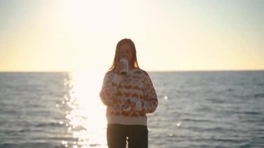 Sıcak süveter ve güneş gözlüğü takan genç ve güzel bir kadın kışın kumsalda dinlenirken termo bardaktan sıcak kahvenin tadını çıkarıyor. Sevimli çekici kız çay içiyor ve okyanus kenarında güneşleniyor..