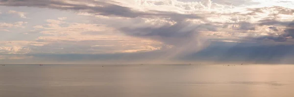 Panorama of stormy sky with sun rays piercing the clouds, flat sea at dawn, minimalist photo and mystery mood.