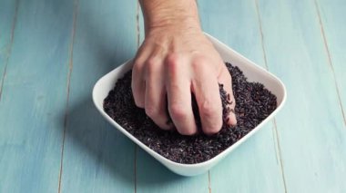 Adam bir kasede siyah pirinç yakalar ve avucunu açar. Sonra da pirinci kasenin içine atar. Mavi arka planda.