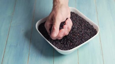 Adam bir kasede siyah pirinç yakalar ve avucunu açar, bir adamın parmakları, beyaz bir kaseye düşen bir avuç siyah pirinç tanelerini hareket ettirir, mavi arka plan..