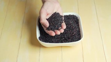 İnsan parmakları, elinde bir avuç siyah pirinç tanesiyle beyaz bir kâse ve sarı arka planda tanınmaz hale gelir..