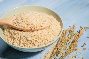 Bir kase kahverengi pirinç, bir kaşık dolusu pirincin ve pirincin yanındaki mavi masaya konur. Çiğ pirinç kaseye yayılır ve kaşık çiğ pirinç ile doldurulur.