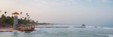 Dominik Cumhuriyeti Bayahibe plajı, bulutlu bir günde, harika dalgalar, engebeli deniz feneri ve ahşap iskeleli romantik deniz feneri.