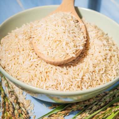 Kahverengi pirinçle dolu yeşil bir kasenin önü, pirinç dolu bir tahta kaşığın üzerinde dinleniyor, hemen yanında bir pirinç kulağı var.