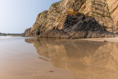 Arka planda kayalık bir uçurum olan bir sahil. Su sakin ve uçurumun yansıması görülebilir.