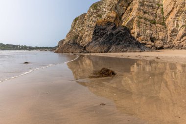 Arka planda kayalık bir uçurum olan bir sahil. Su sakin ve uçurumun yansıması görülebilir.