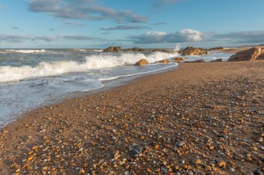Dağınık çakıl taşları ve deniz kabuklarıyla geniş bir kumsal uzanır. Hafif dalgalar, öğleden sonra saatlerinde kısmen bulutlu bir gökyüzünün altında kıyıya çarpar. Plage de l 'Aubraie, Les Sables d' Olonne, Vendee, Fransa, Europa.