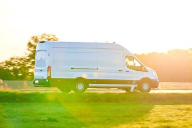 Beyaz minibüs köy yolunda hızlı gidiyor. Teslimat kamyonu sonbahar mevsiminde gün batımında sürüyor.