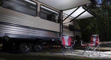 Kamp karavanı ormanlık bir kamp alanına kurulmuş, tenteleri açık ve altında sandalyeler var.
