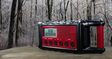 Güneş enerjili bir hava durumu radyosunun arkasında buzla kaplı bir orman ve LCD ekranında bir buz fırtınası görüntüsü var..