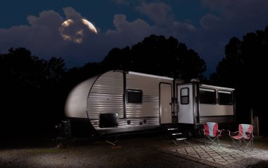 Travel trailer and camp all set up in a thick forest with the moon peeking through the clouds in the sky behind. clipart