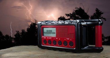 Önplanda kırmızı kablosuz hava durumu radyosuyla arka planda yıldırım çarpması ön plandaki LCD ekranında şiddetli bir fırtına uyarısı gösteriyor..