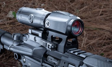 AR-15 'in arkasında kırmızı bir optik monte ve büyüteç var. Gündüz vakti çam iğneleriyle kaplı bir ormanda kullanılmaya hazır.. 