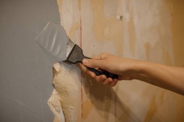 Metal kazıyıcı kullanan, eski duvar kağıtlarını söken ya da yenilenirken desenli bir duvardan boya çıkaran bir insan eli.